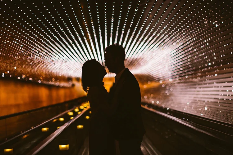 Silhouetted couple kissing inside a modern tunnel with illuminated rows of small lights forming a pattern on the ceiling.