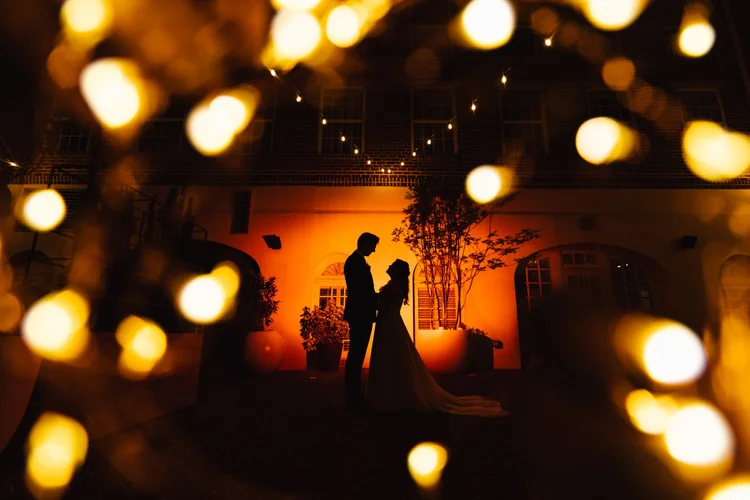 Silhouette of a couple at their wedding or romantic event, standing close and holding hands, illuminated by warm orange lighting, viewed through blurred string lights.
