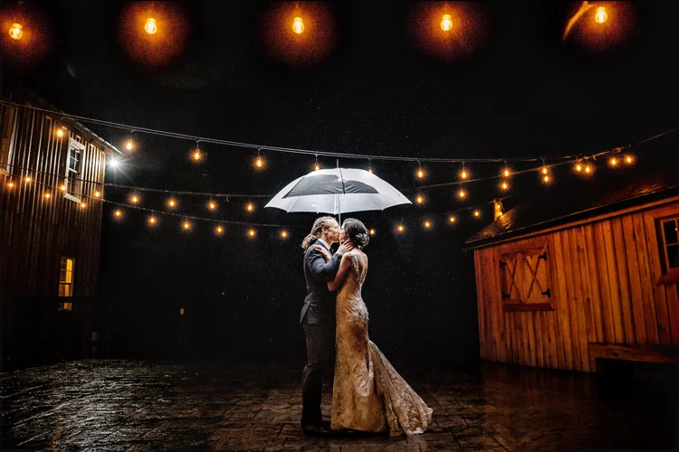 A couple shares a kiss under an umbrella on a rainy night, illuminated by string lights, with wooden buildings in the background.