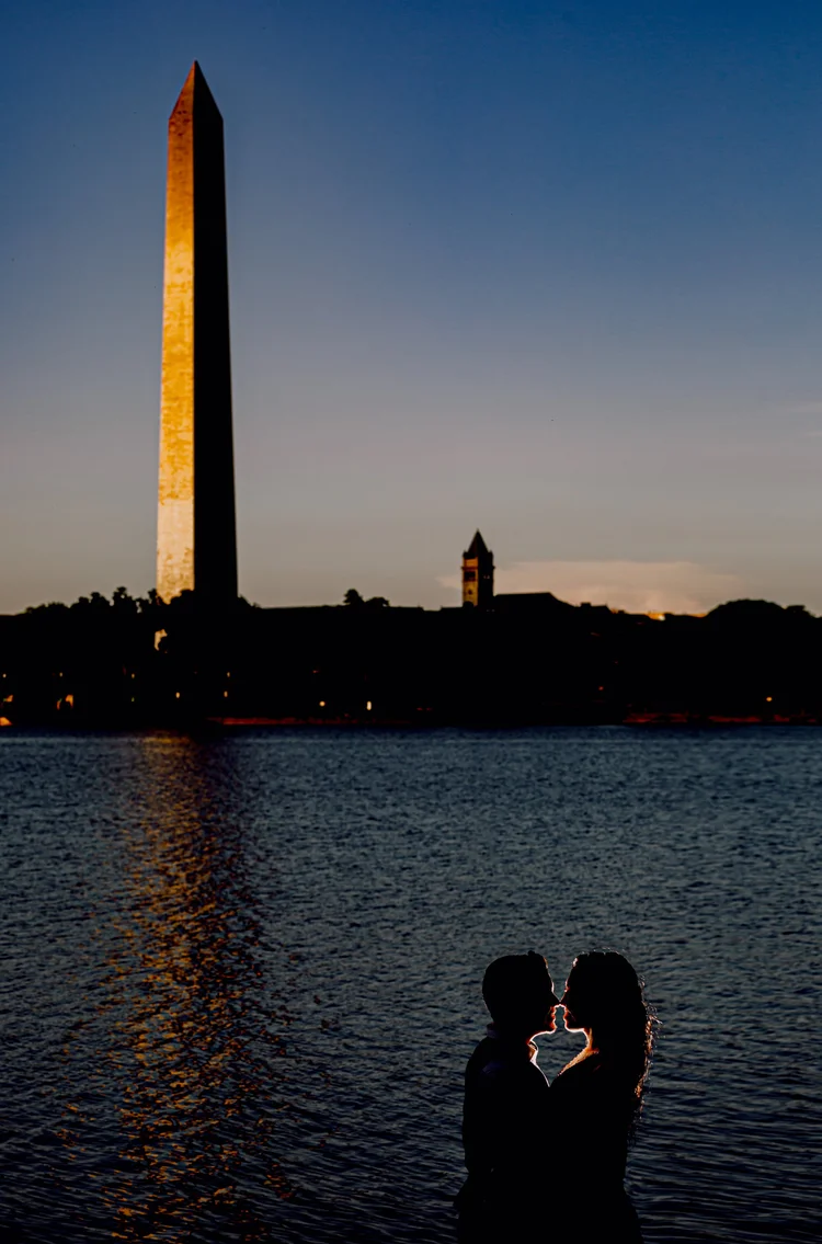 Silhouette of a couple by the water with the Washington Monument and a monument with a clock tower in the background during sunset.
