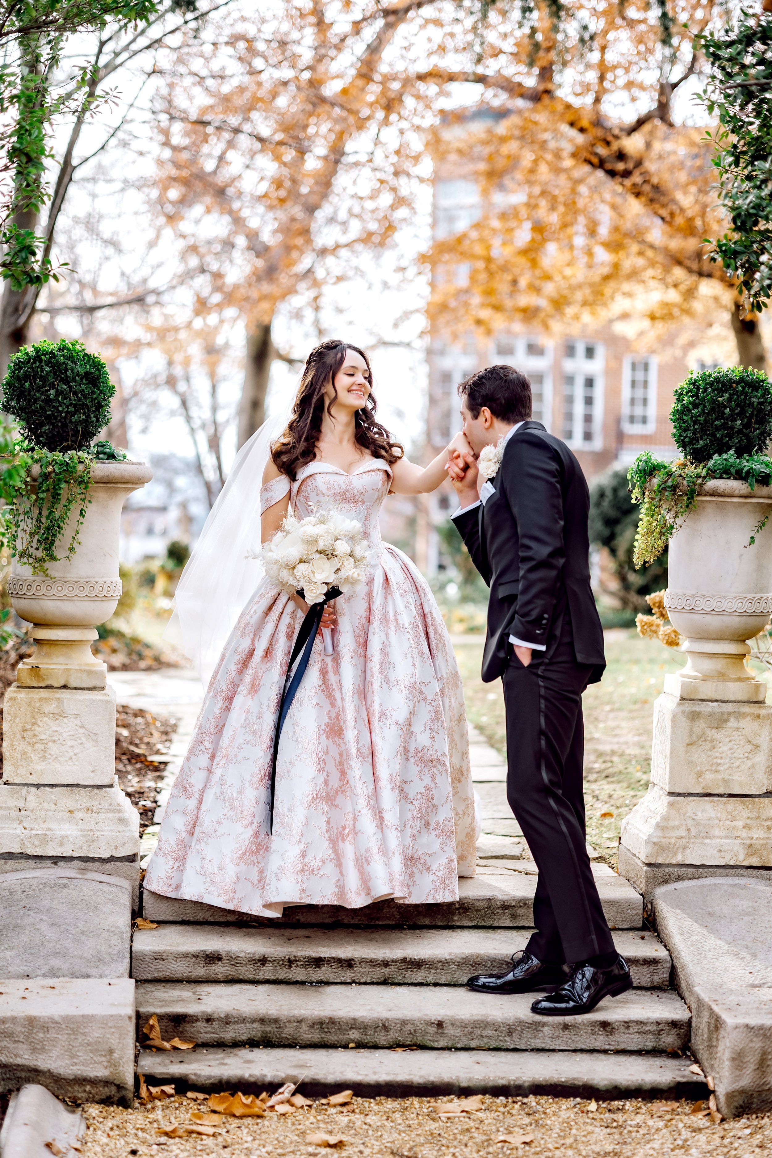 A bride and groom in wedding attire are outdoors on a stone staircase, with the bride smiling as he kisses her hand. The bride is wearing a vintage-style off-shoulder wedding gown with a full skirt, holding a bouquet of white roses. The groom is dres