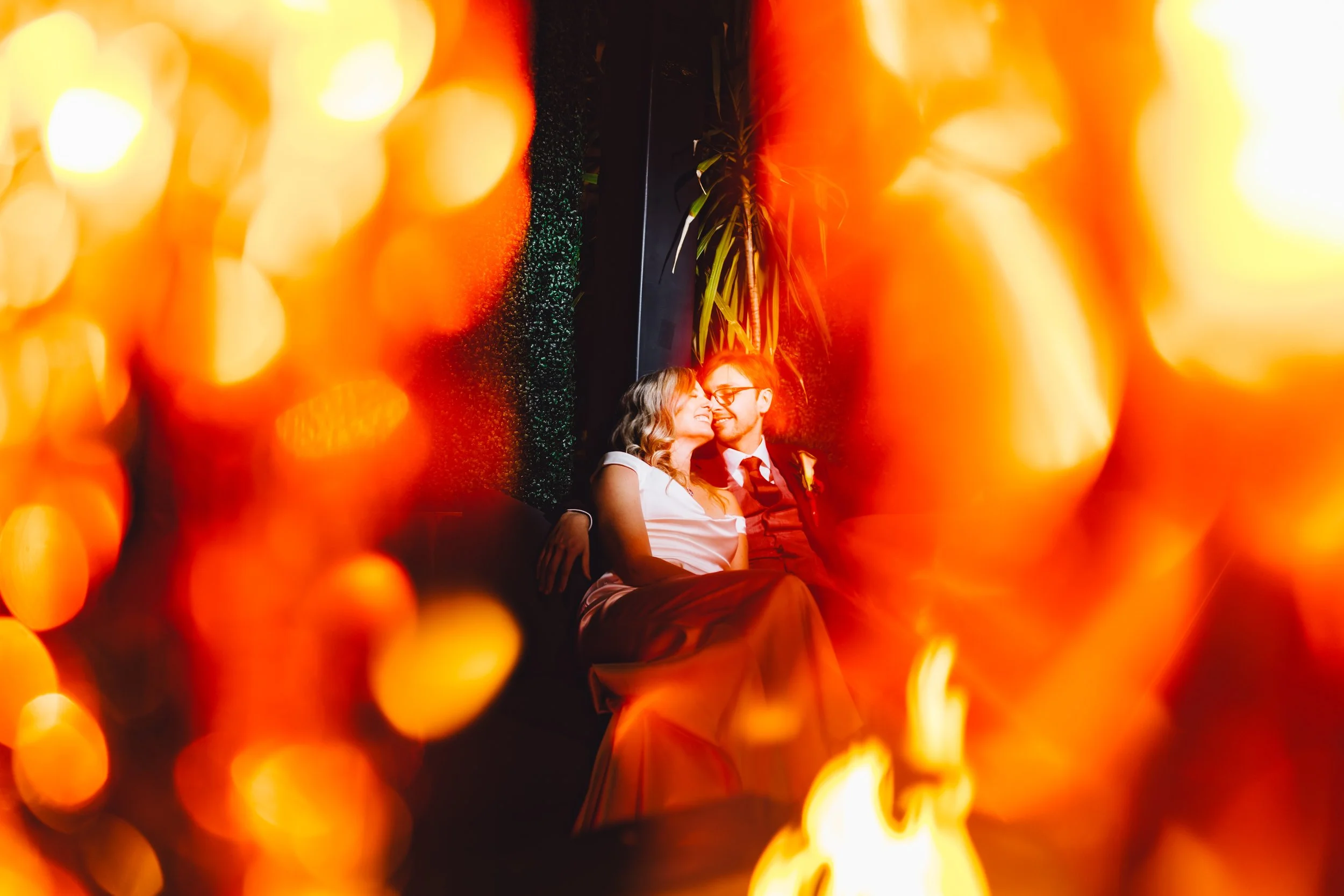 A couple sitting close together at night, smiling and looking into each other's eyes. The woman has wavy hair and is wearing a white sleeveless dress. The man is wearing glasses, a dress shirt, and a suit. They are surrounded by warm, orange bokeh li
