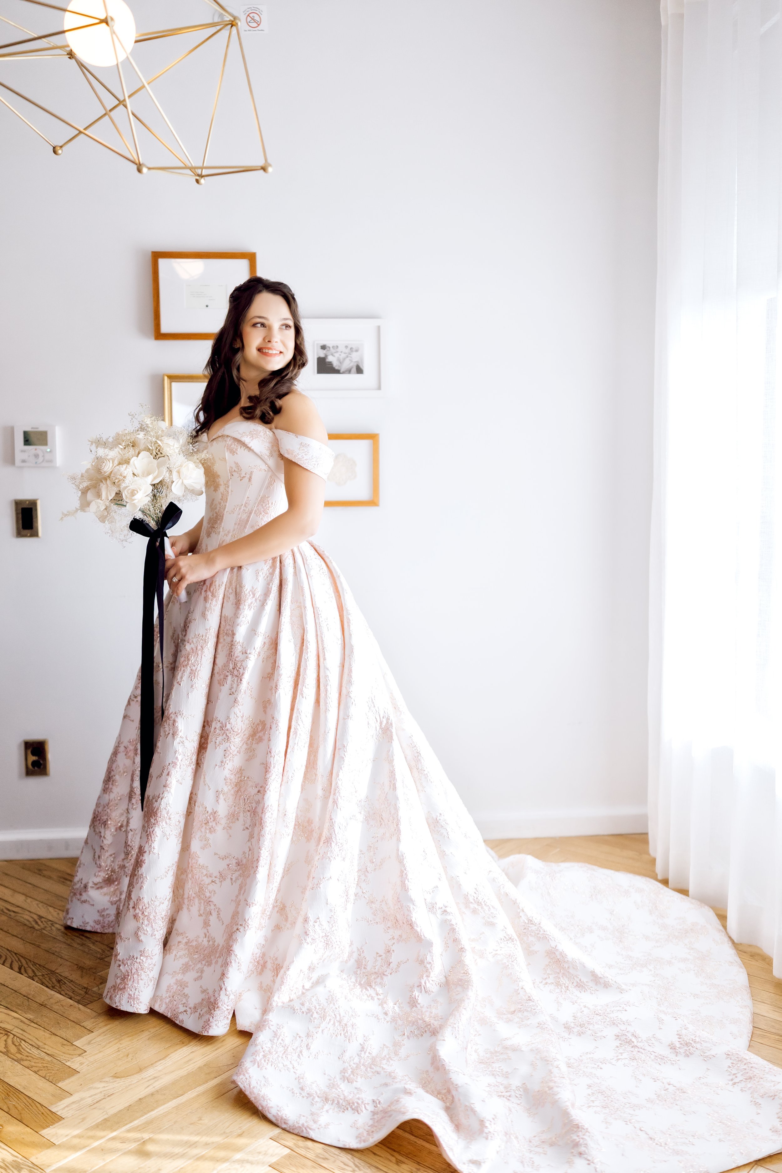 A woman in a wedding dress holding a bouquet of white flowers standing indoors near a window with sheer curtains. The room has hardwood floors, framed pictures on the wall, and a modern geometric light fixture hanging from the ceiling.