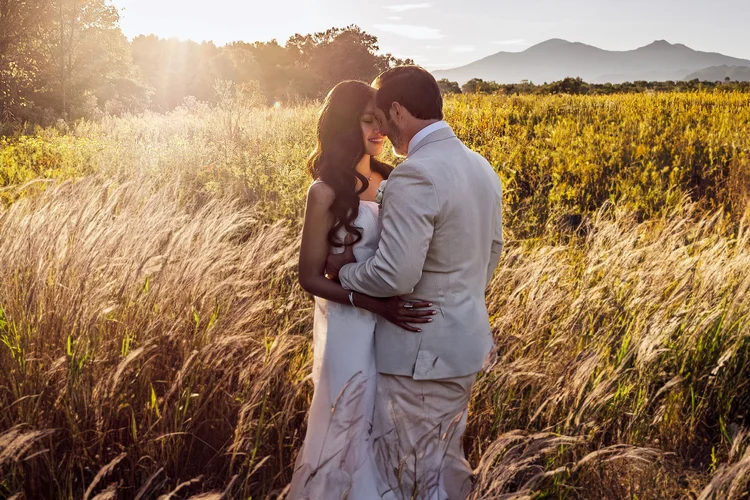 A couple dressed in wedding attire standing close together in a field of tall grass, with mountains and trees in the background, illuminated by sunset light.