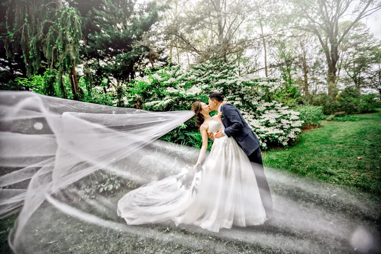A bride and groom kissing outdoors, with the bride in a white gown and veil, and the groom in a dark suit, surrounded by green trees and bushes.