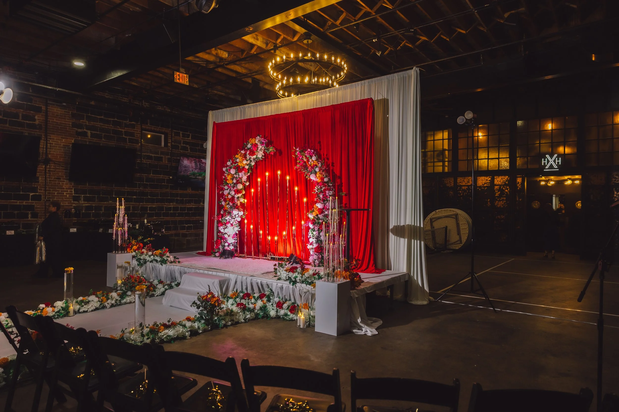 A decorated stage with a red curtain, floral arrangements, candles, and string lights, set for a wedding or special event inside a dimly lit venue.