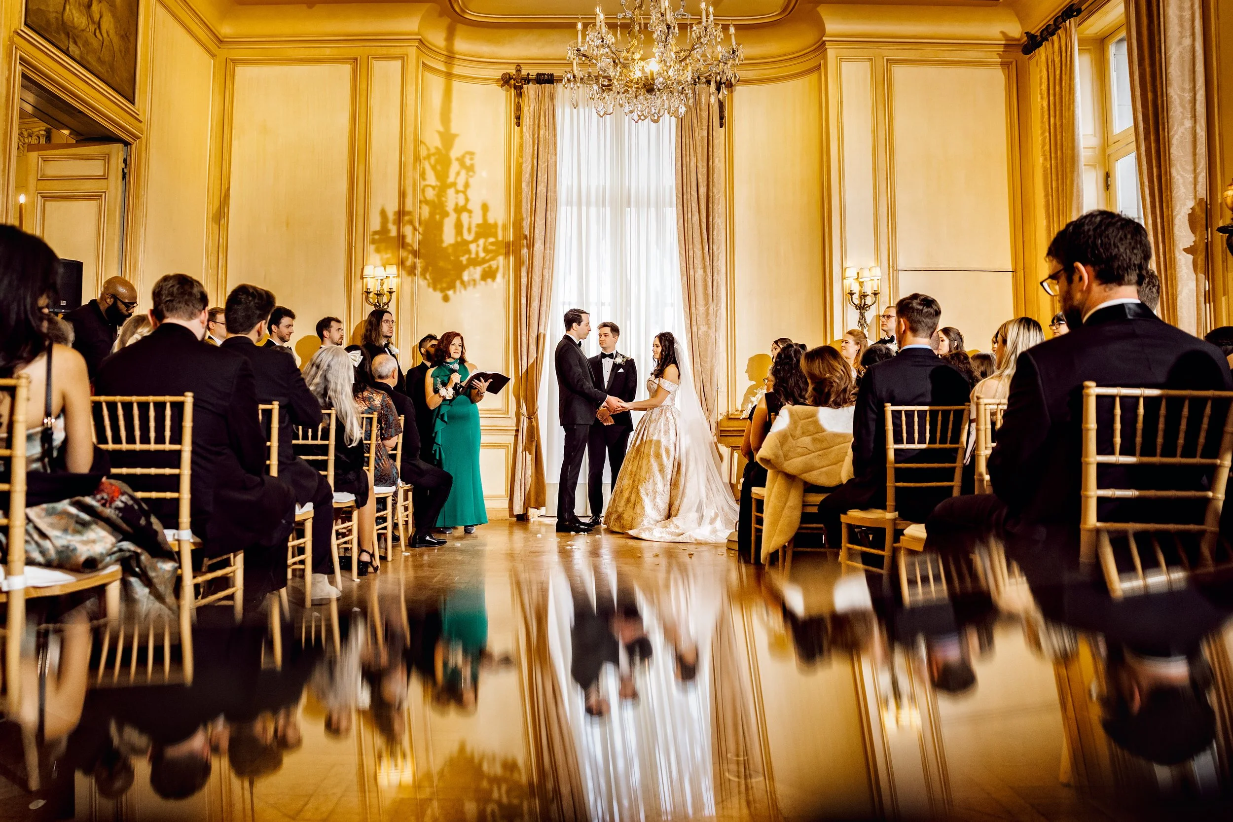 A wedding ceremony in an elegant, grand room with gold walls, large window, chandelier, and guests seated on Gold chairs.