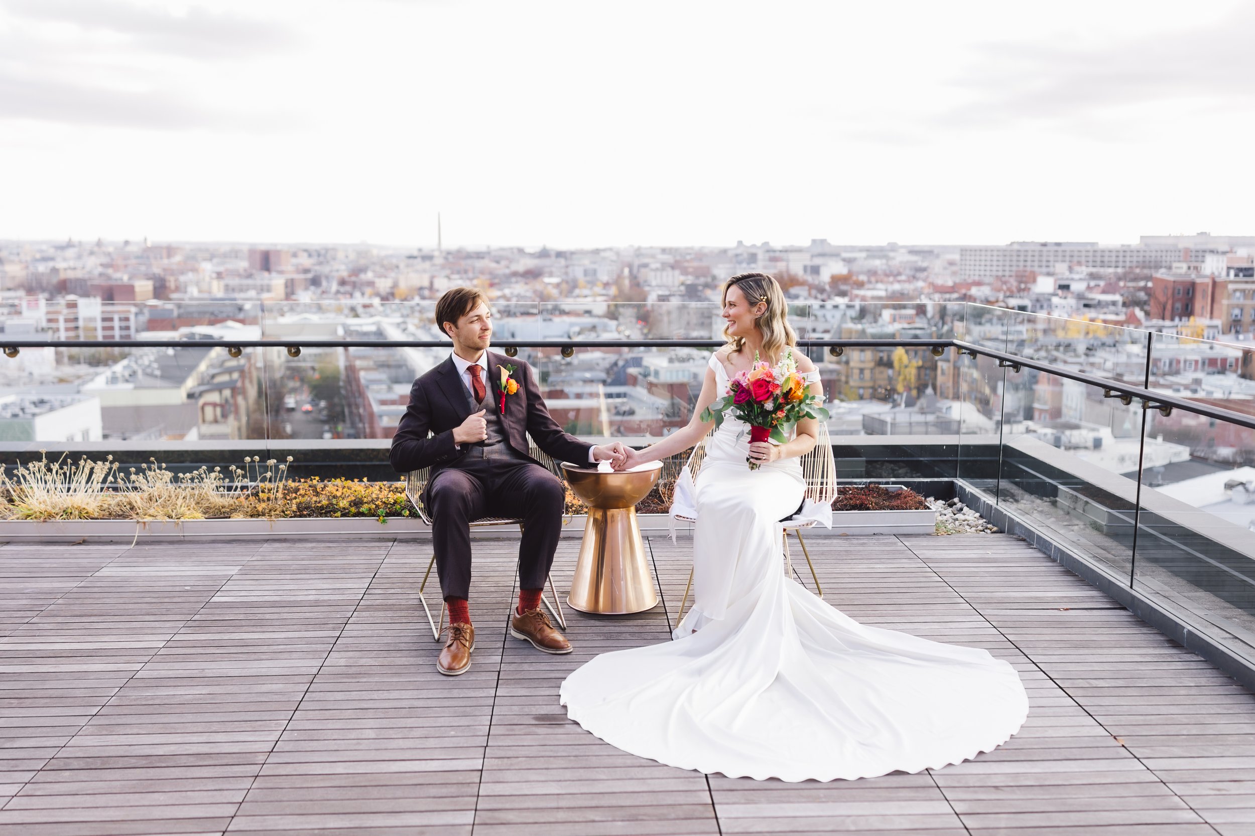 A bride and groom sitting on a rooftop, holding hands, with a city skyline in the background. The bride wears a white wedding dress and holds a bouquet of flowers, while the groom wears a dark suit with a red boutonniere.