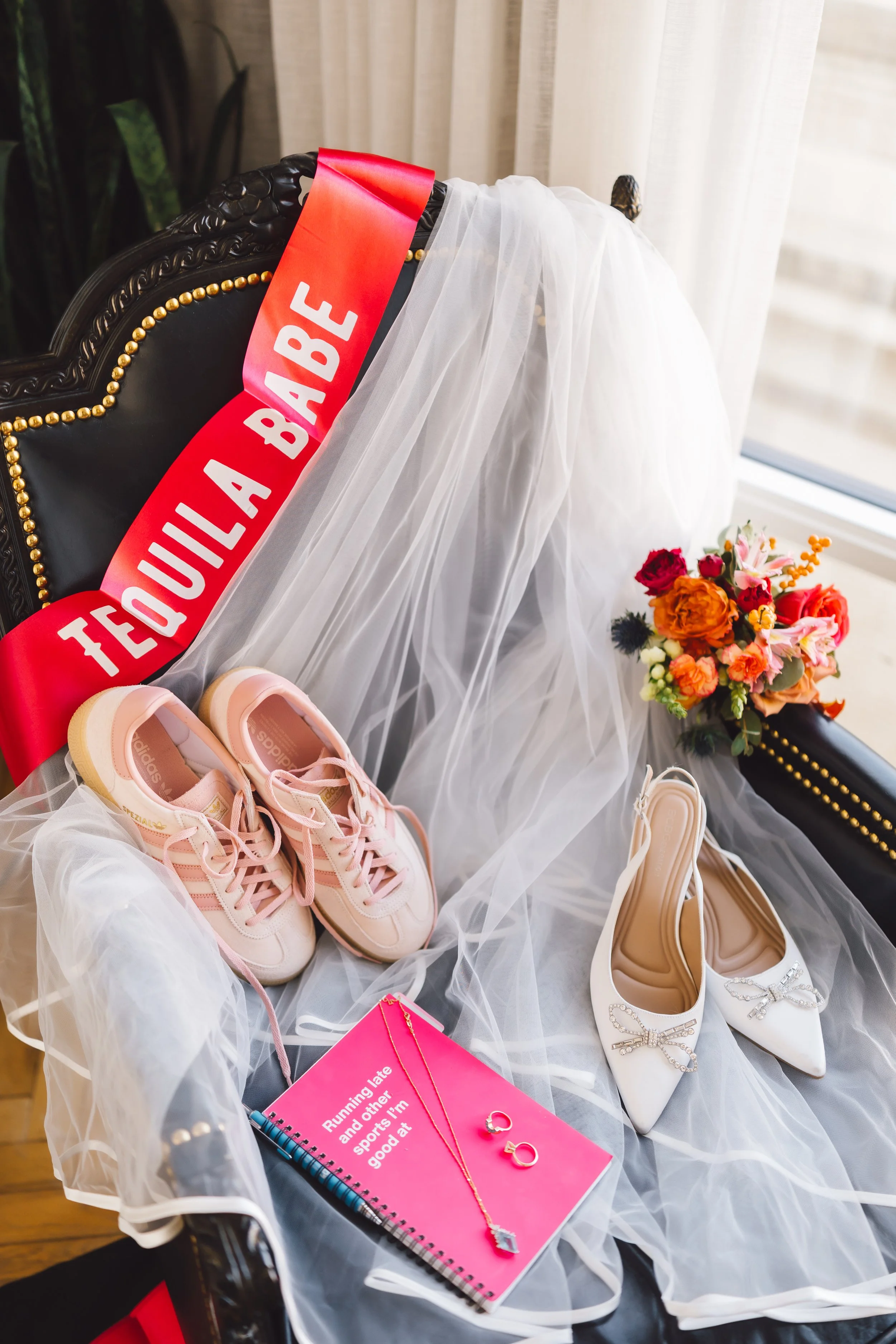 A black chair decorated with a white veil, pink sneakers, white high heels with bows, a pink notebook, jewelry, and a bouquet of flowers, with a red sash reading 'FEOUILA A BAILE' draped over the chair, situated by a window.