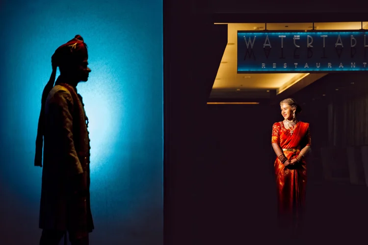 A woman in a red traditional dress smiling and looking at a man in a traditional outfit standing to the left, in front of a blue-lit wall with a restaurant sign overhead.