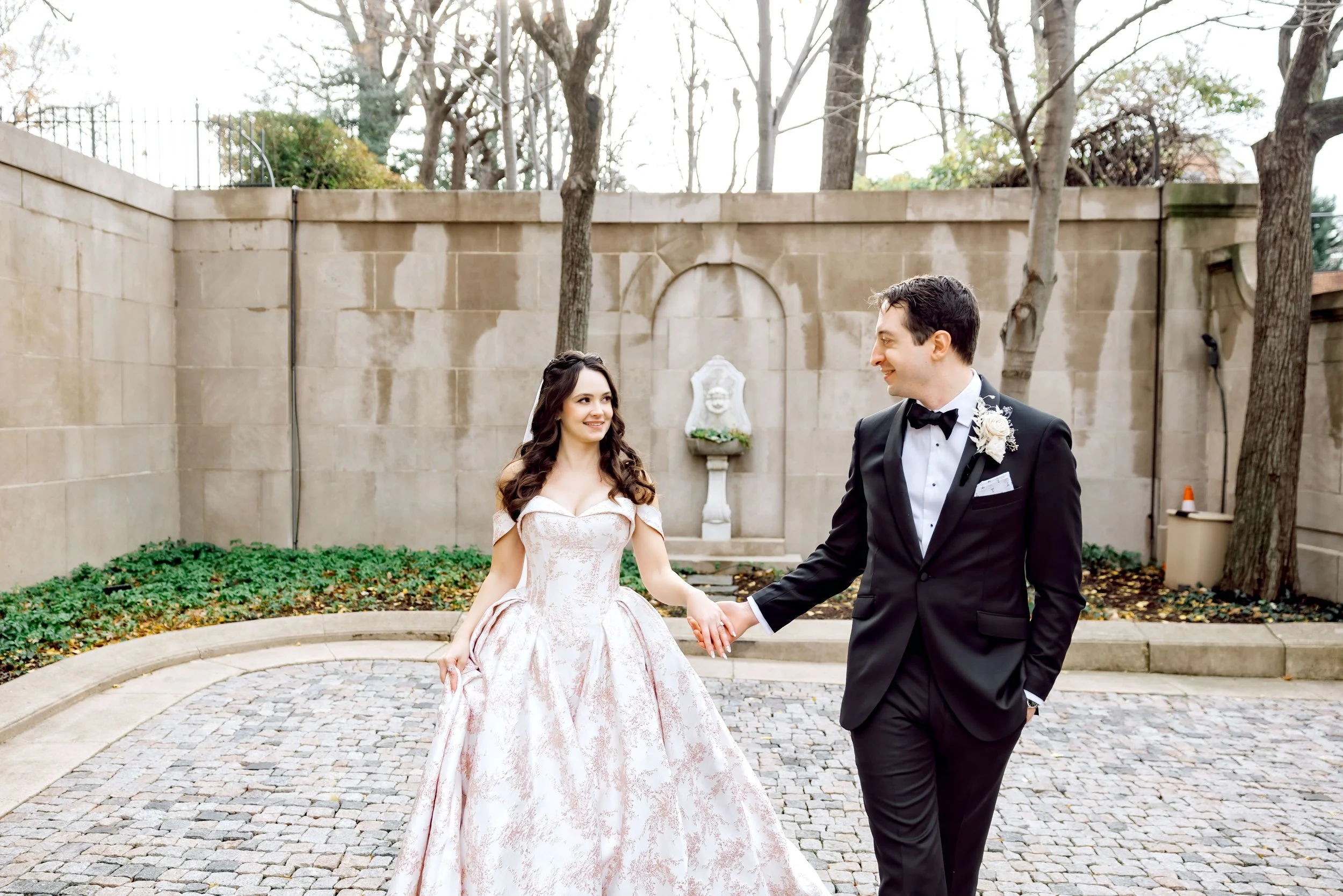 A bride and groom holding hands outside in a courtyard, with stone walls and trees in the background during daytime.