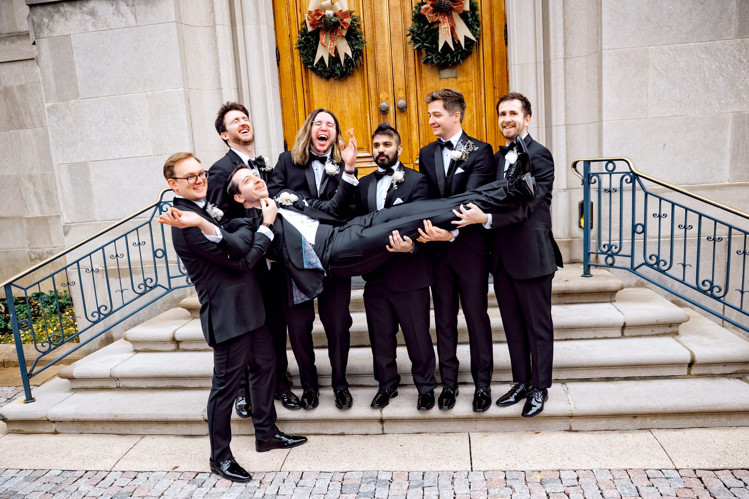 A group of seven men dressed in black tuxedos with bow ties, holding a man dressed in the same attire horizontally in front of a wooden door adorned with holiday wreaths. The men are smiling and laughing outside a building with stone steps and railin