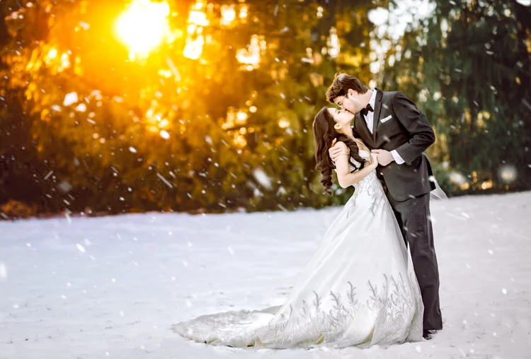 A newlywed couple kisses in a snowy outdoor setting at sunset, with trees in the background.
