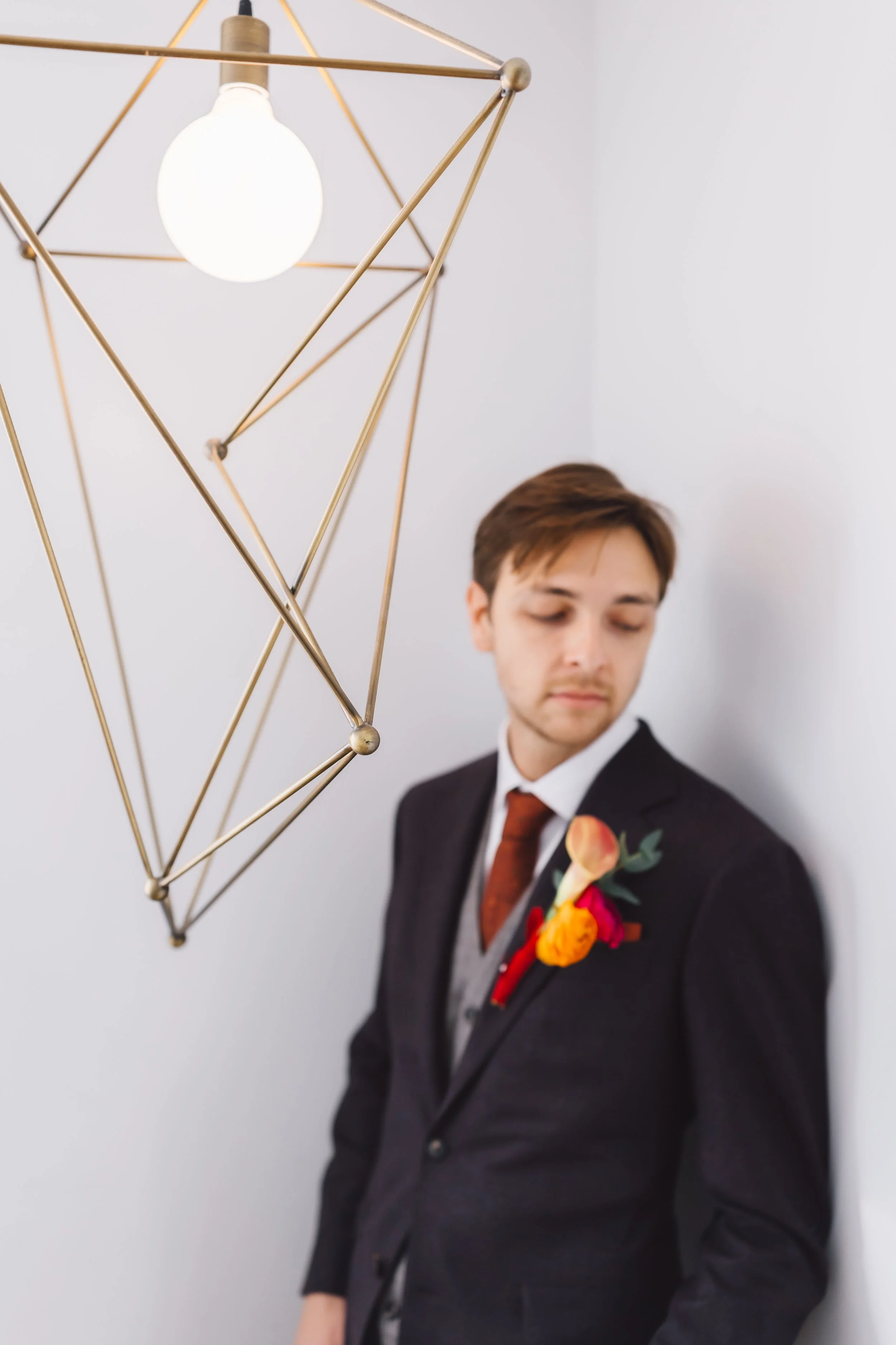 A young man in a suit and tie leaning against a white wall, with a geometric gold light fixture and a bright white light bulb overhead.