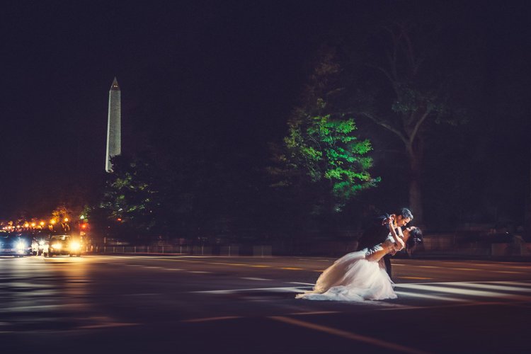 A couple dressed in wedding attire sharing a dance on a city street at night, with the Washington Monument visible in the background and a line of cars with headlights on.