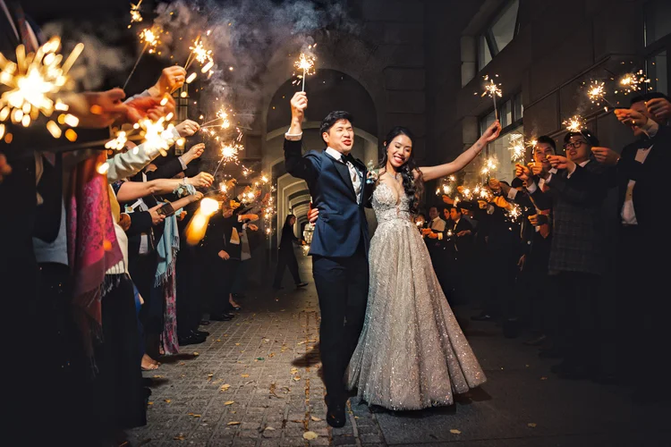 A newlywed couple in wedding attire walking through a corridor surrounded by guests holding sparklers during celebration.
