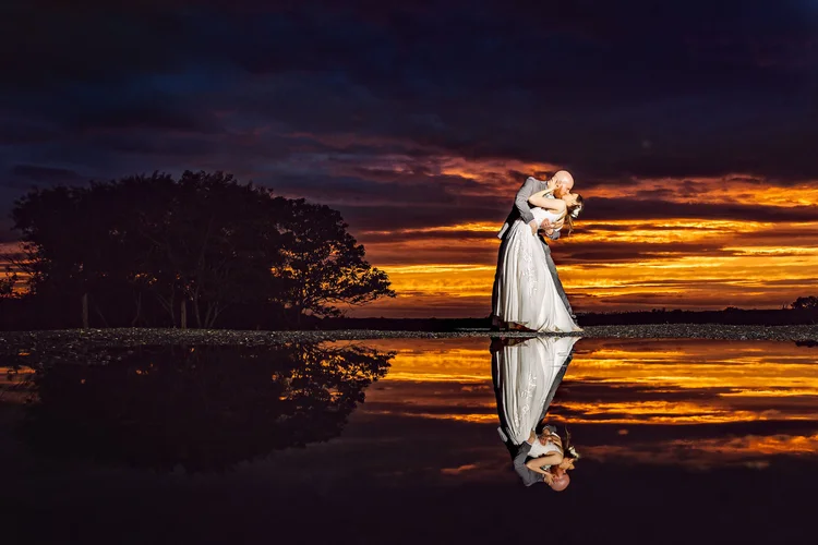 A couple in wedding attire sharing a kiss during sunset with a reflection of their figures on a nearby body of water.