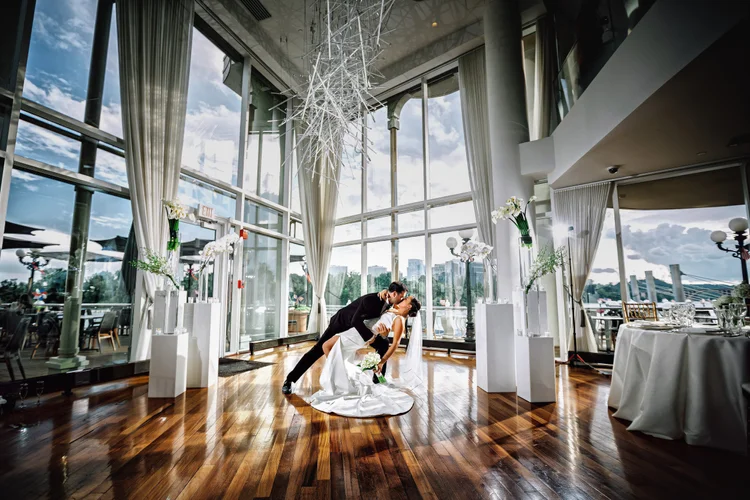 A newlywed couple sharing their first dance inside a glass-walled wedding reception venue with a cityscape view, decorated with white flowers and elegant furnishings.