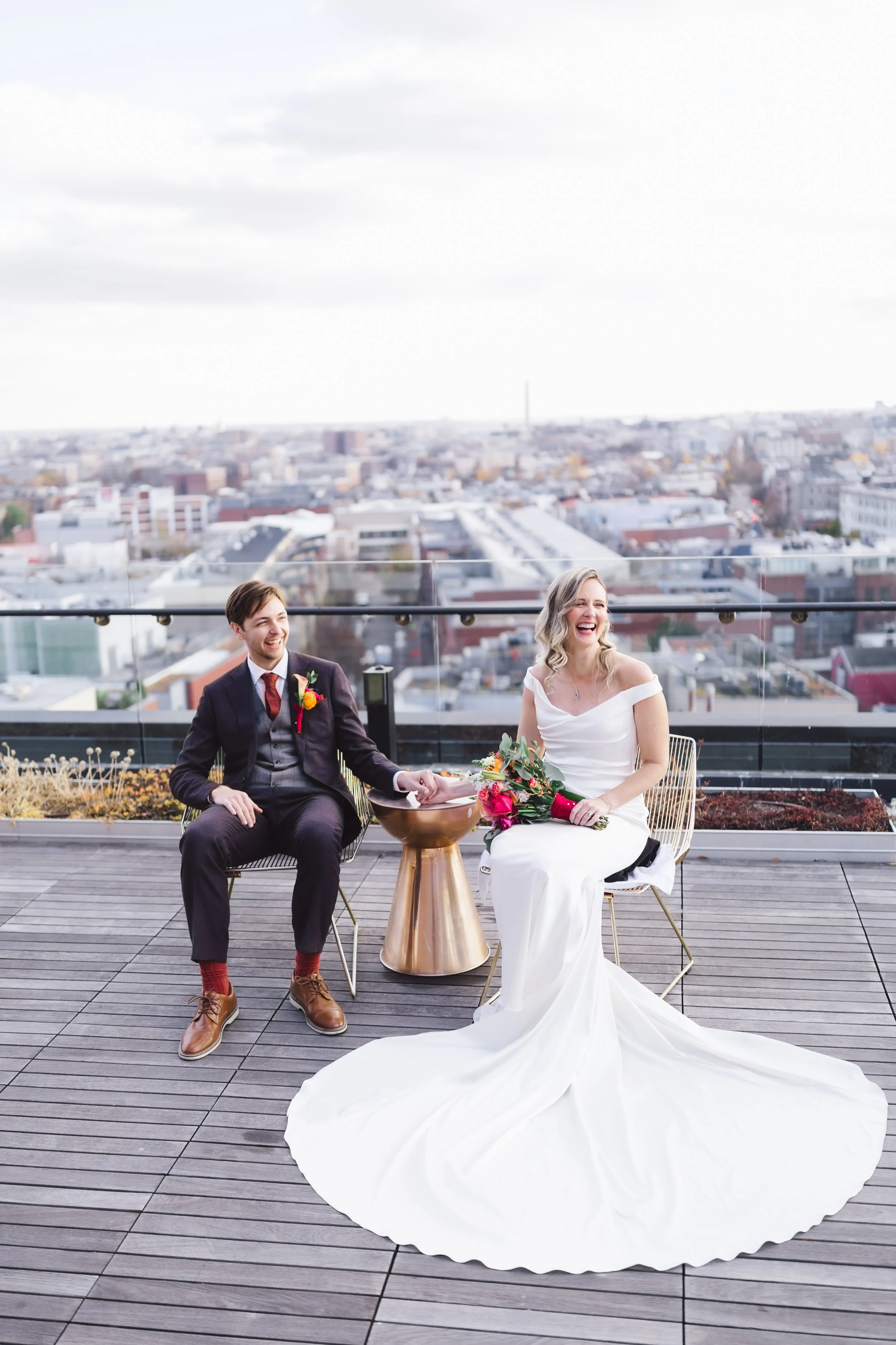 A newlywed couple sitting on a rooftop terrace, enjoying the city view, with the bride in a white wedding dress holding a bouquet of flowers, and the groom in a suit, both smiling and laughing.