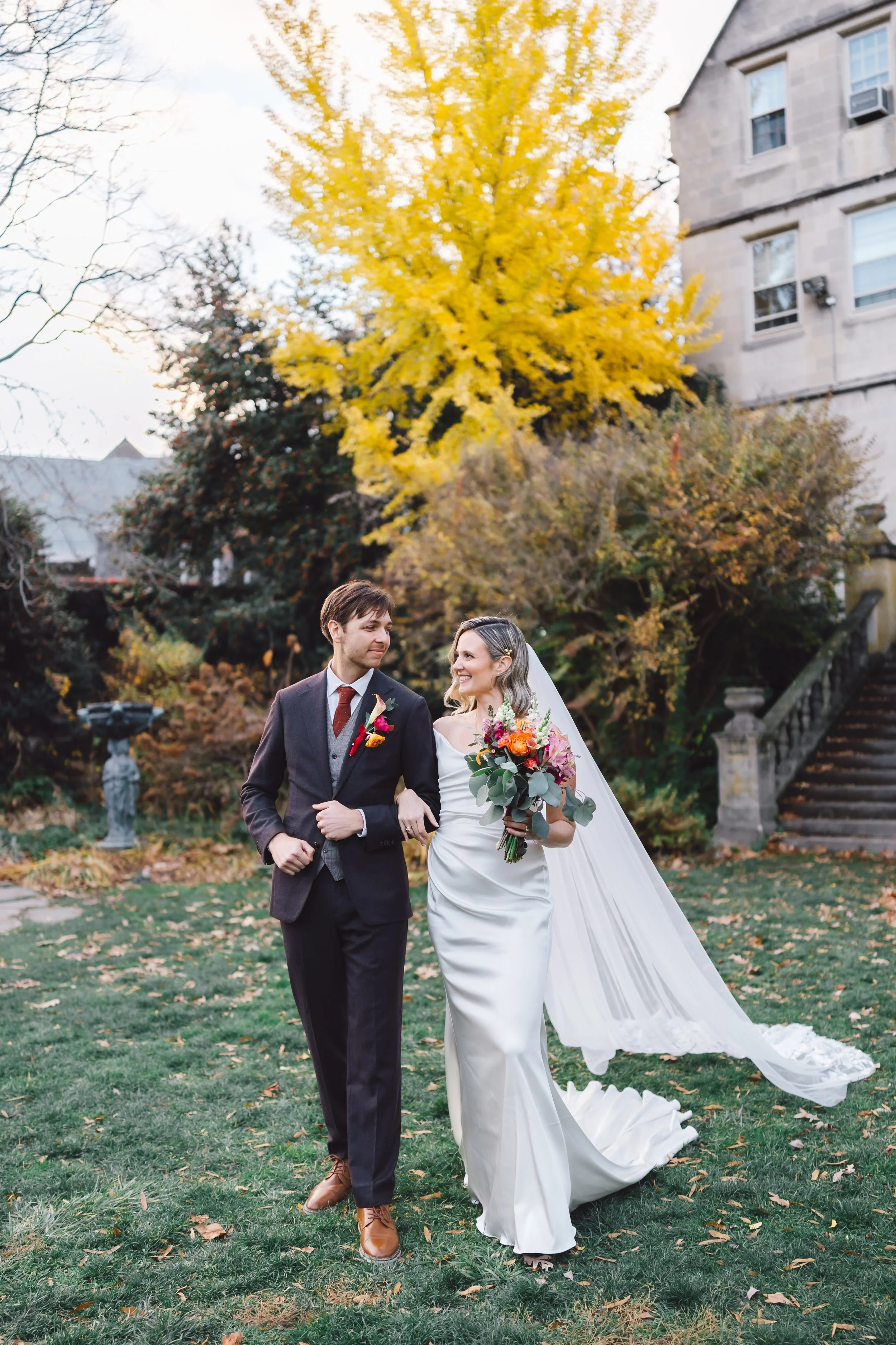A bride in a white wedding gown and a groom in a dark suit walk outdoors, the bride holding a colorful bouquet, with autumn trees and a building in the background.