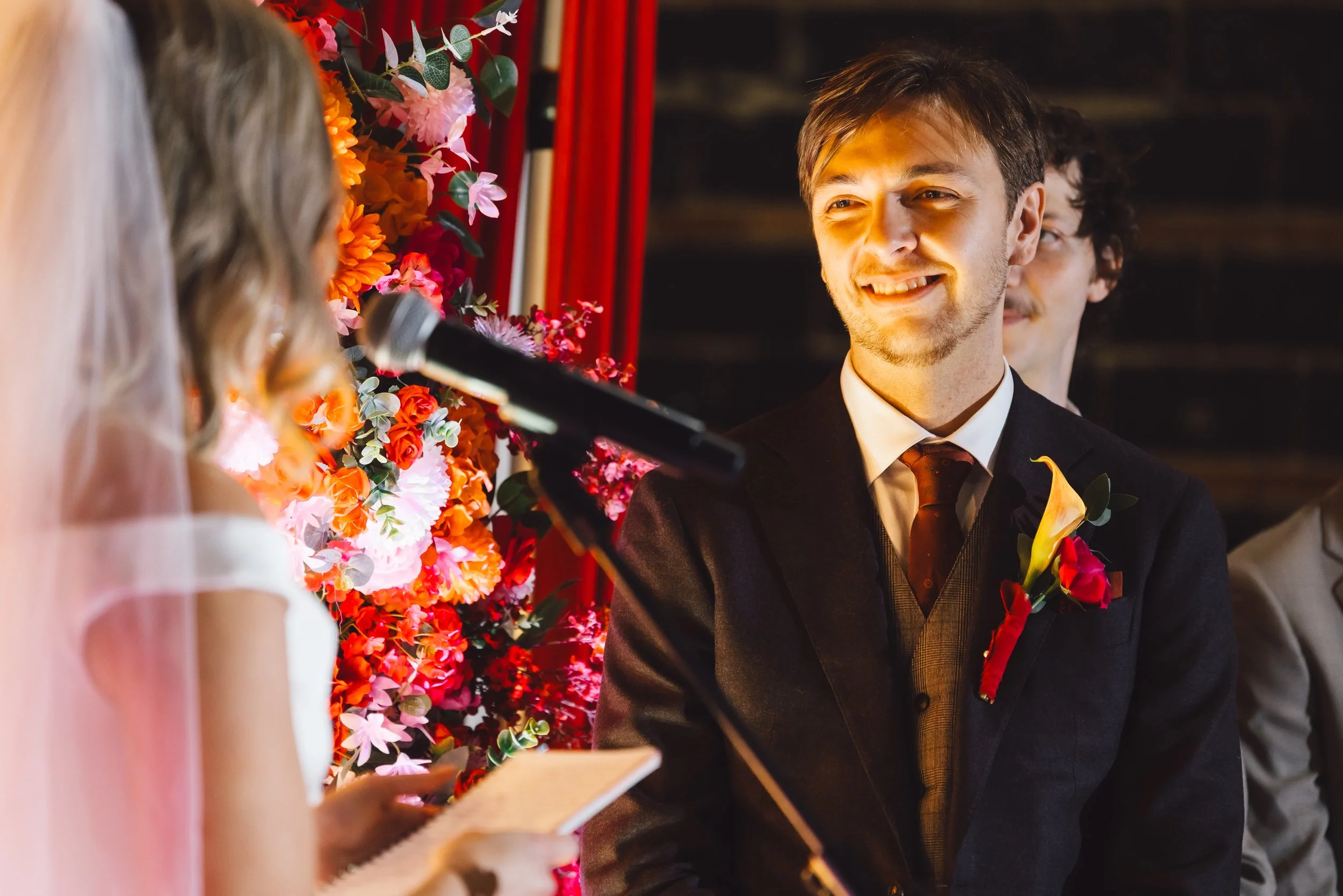Man in a black suit with boutonniere smiling at bride during wedding ceremony, with floral backdrop, woman reading vows, and man with glasses in background.