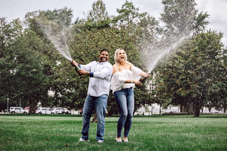 A joyful couple celebrating outdoors on a grassy field, each holding a champagne bottle and spraying champagne into the air.