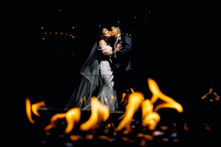 A couple in wedding attire share a kiss at night with fire in the foreground and string lights in the background.