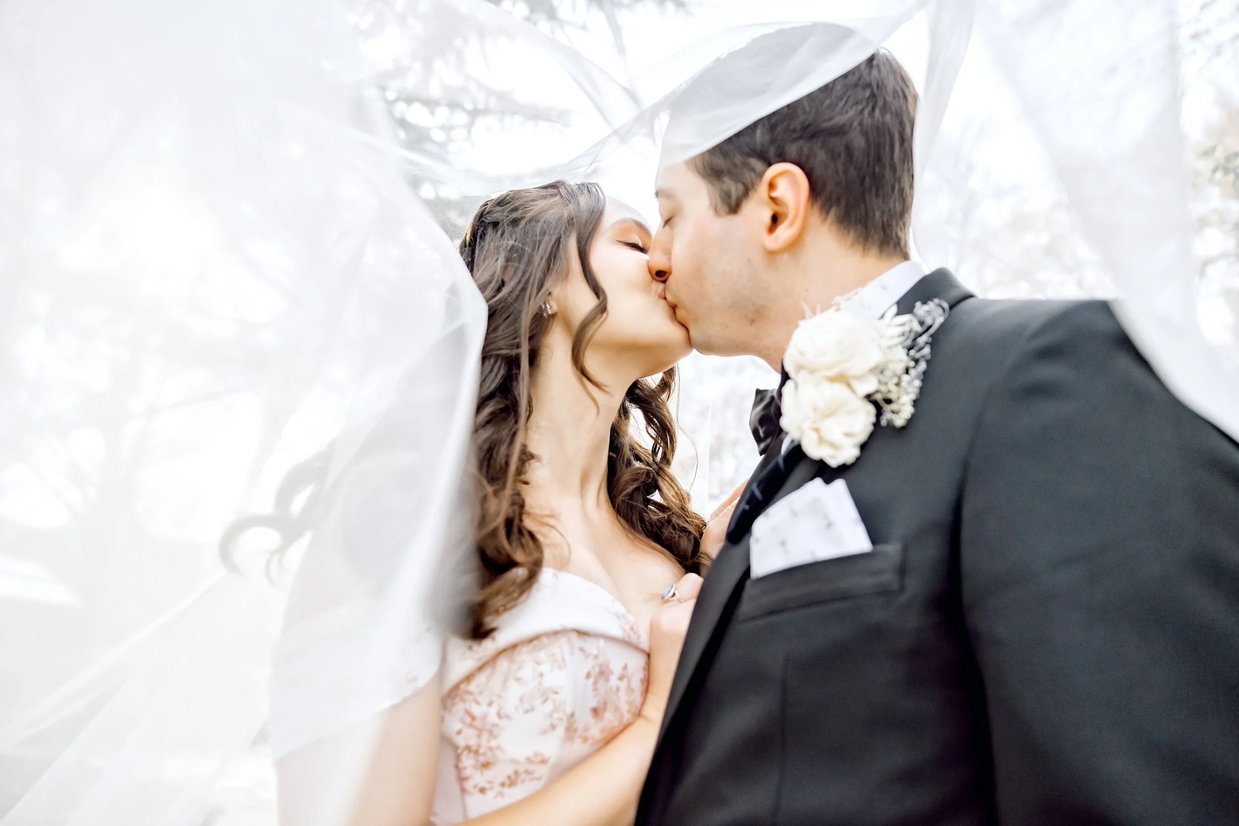 A bride and groom sharing a kiss under a veil during their wedding outdoors.