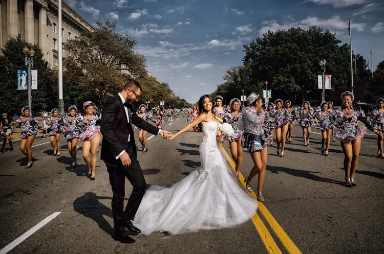 Bride and groom holding hands in the street with cheerleaders dancing around them during a parade or celebration.