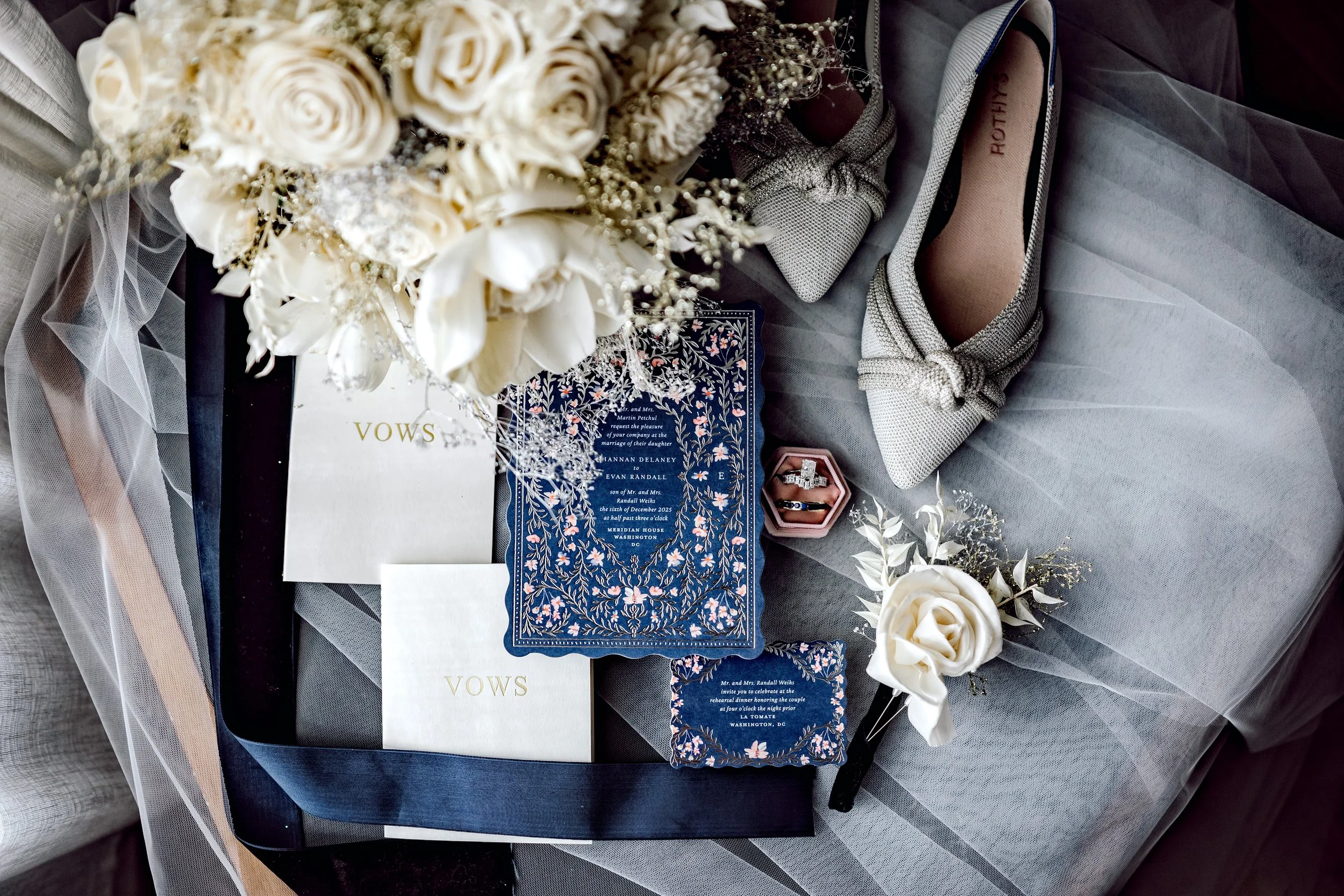 Wedding accessories including vows, bridal bouquet with white roses, wedding rings in a pink box, gray shoes with bows, and decorative paper on a gray fabric surface.