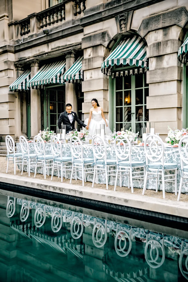 Bride and groom holding hands at an outdoor wedding reception table decorated with floral centerpieces, white chairs, and floral-patterned tablecloths, in front of a historic stone building with green and white striped awnings, and a reflecting pool 