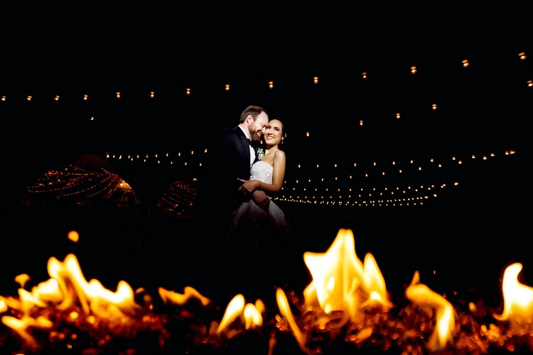 A newlywed couple dancing at night with string lights overhead and a fire in the foreground.
