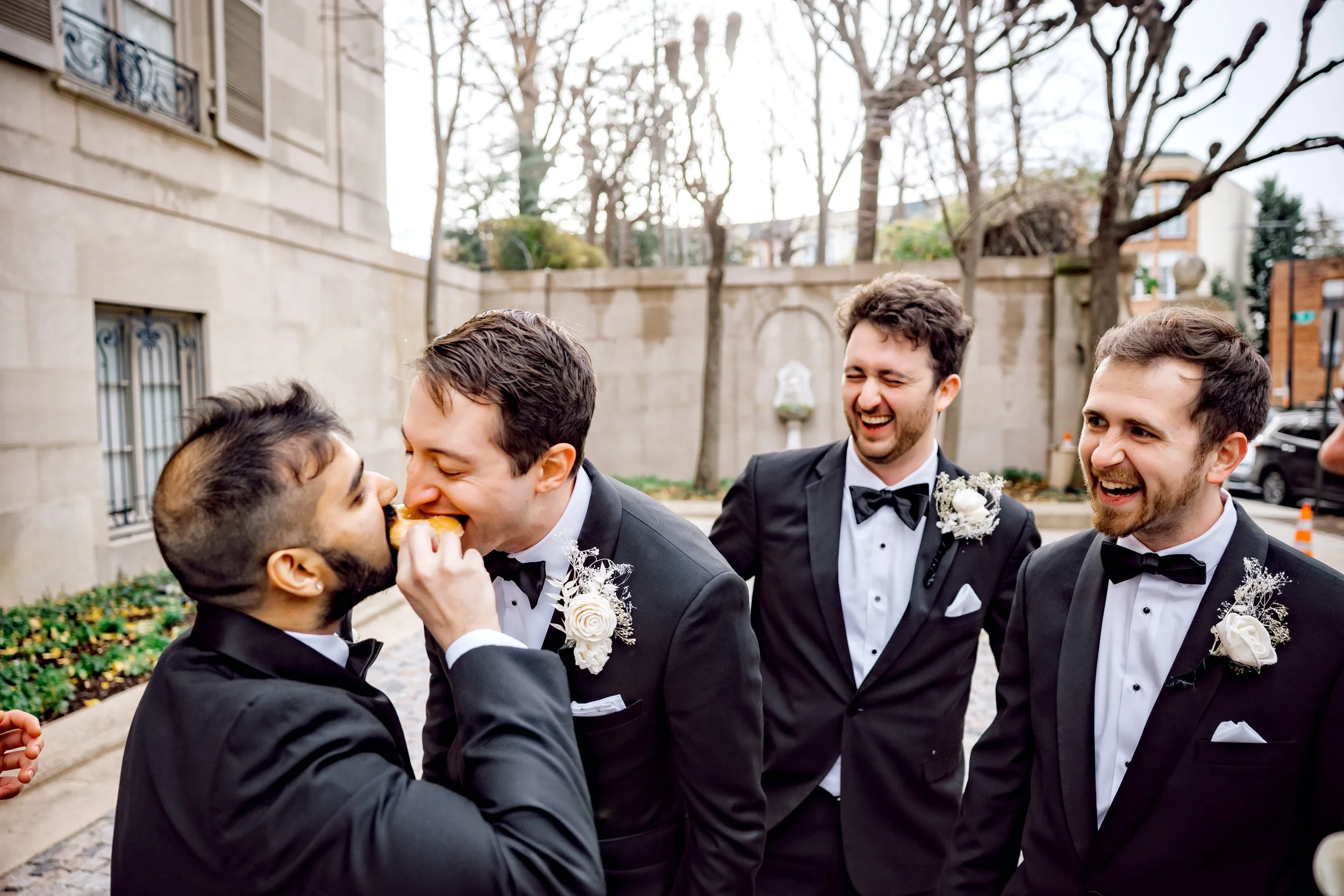 Four men in tuxedos with boutonnières, outdoors in a garden area, one man is being fed cake by another, all are smiling and laughing.