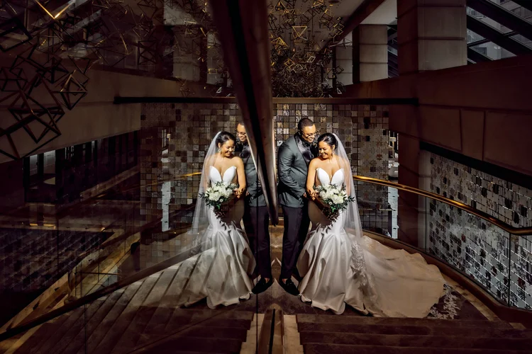 Reflection of a bride and groom on a glass surface, standing on stairs inside a modern building, with the bride in a white wedding gown and the groom in a dark suit, smiling and holding bouquets.