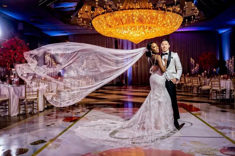 A bride and groom dancing at their wedding reception in a ballroom with a large, ornate chandelier, decorated tables, and floral centerpieces.