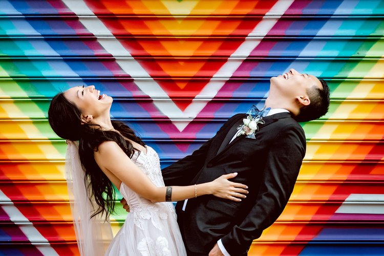 A bride and groom in wedding attire laughing and holding each other in front of a colorful, graffiti-style mural.