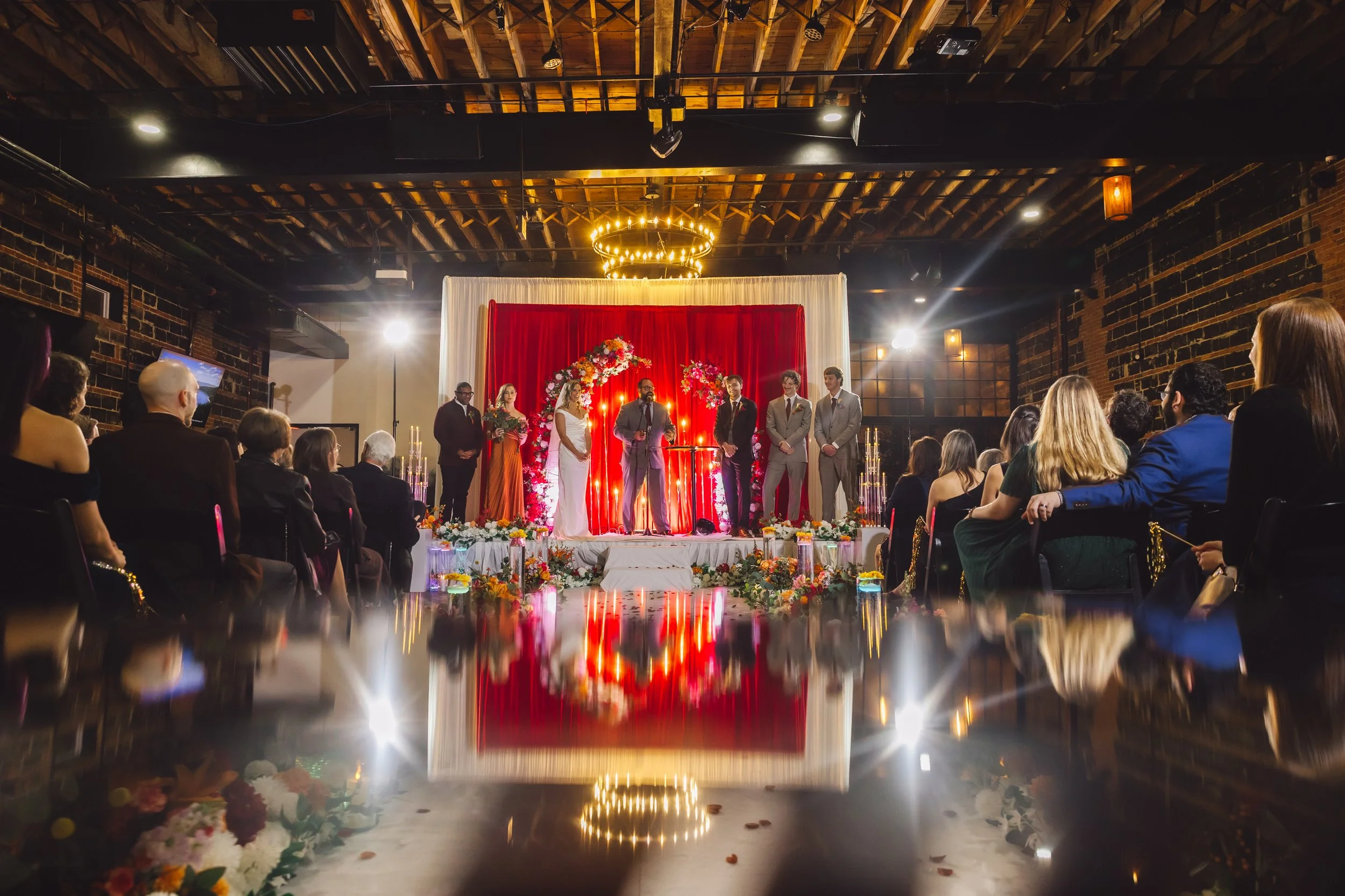 A wedding ceremony with the bride and groom standing on a decorated stage with bridesmaids and groomsmen. The audience is sitting and watching in a dimly lit venue with exposed brick walls and wooden ceiling. The stage has floral arrangements and can