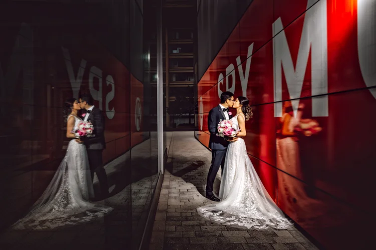 A bride and groom sharing a kiss in an urban setting at night, with the bride holding a bouquet of pink and white flowers and a large red wall with white text in the background. Their reflection is visible in the glass wall next to them.