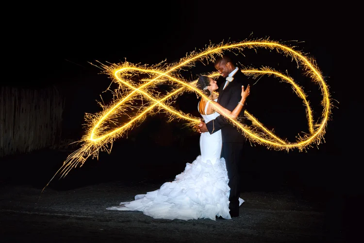 A newlywed couple dancing at night with sparklers creating light trails around them.