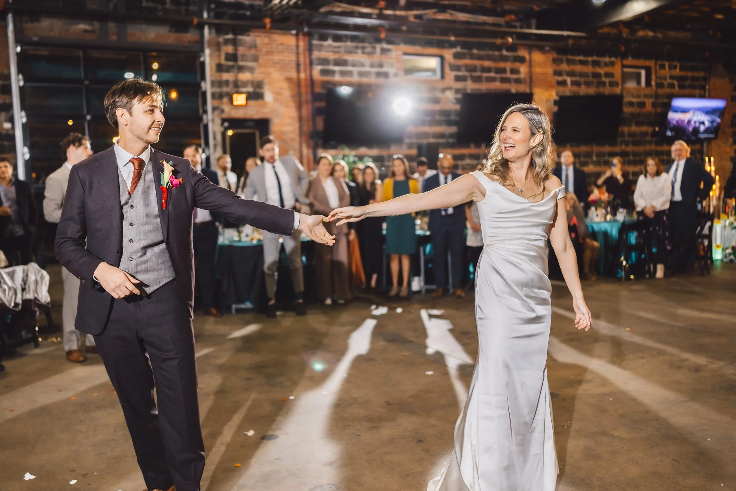 A bride and groom dancing at a wedding reception, with guests watching in the background. The bride is wearing a satin white gown, and the groom a dark suit with a vest and tie. The celebration is in a rustic, brick-walled venue.