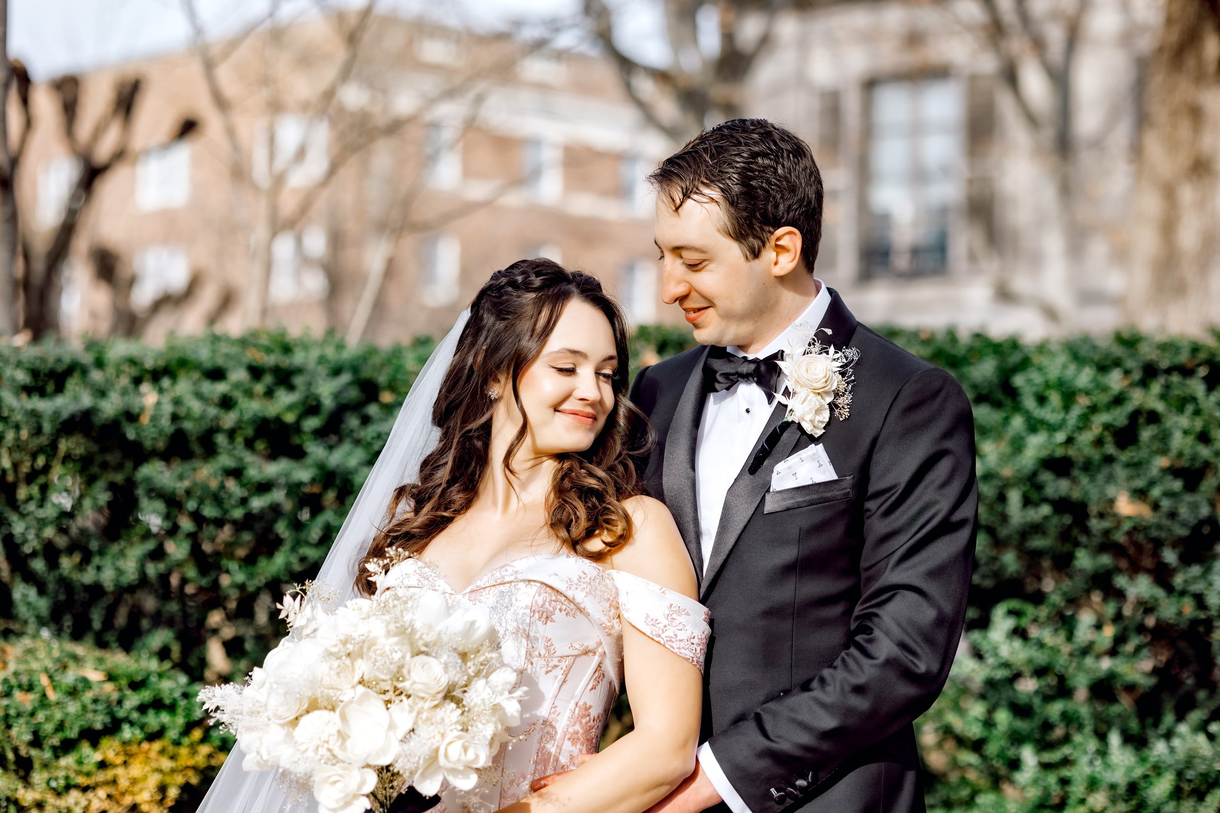 Bride and groom on their wedding day, standing outdoors with greenery and buildings in the background, smiling at each other.