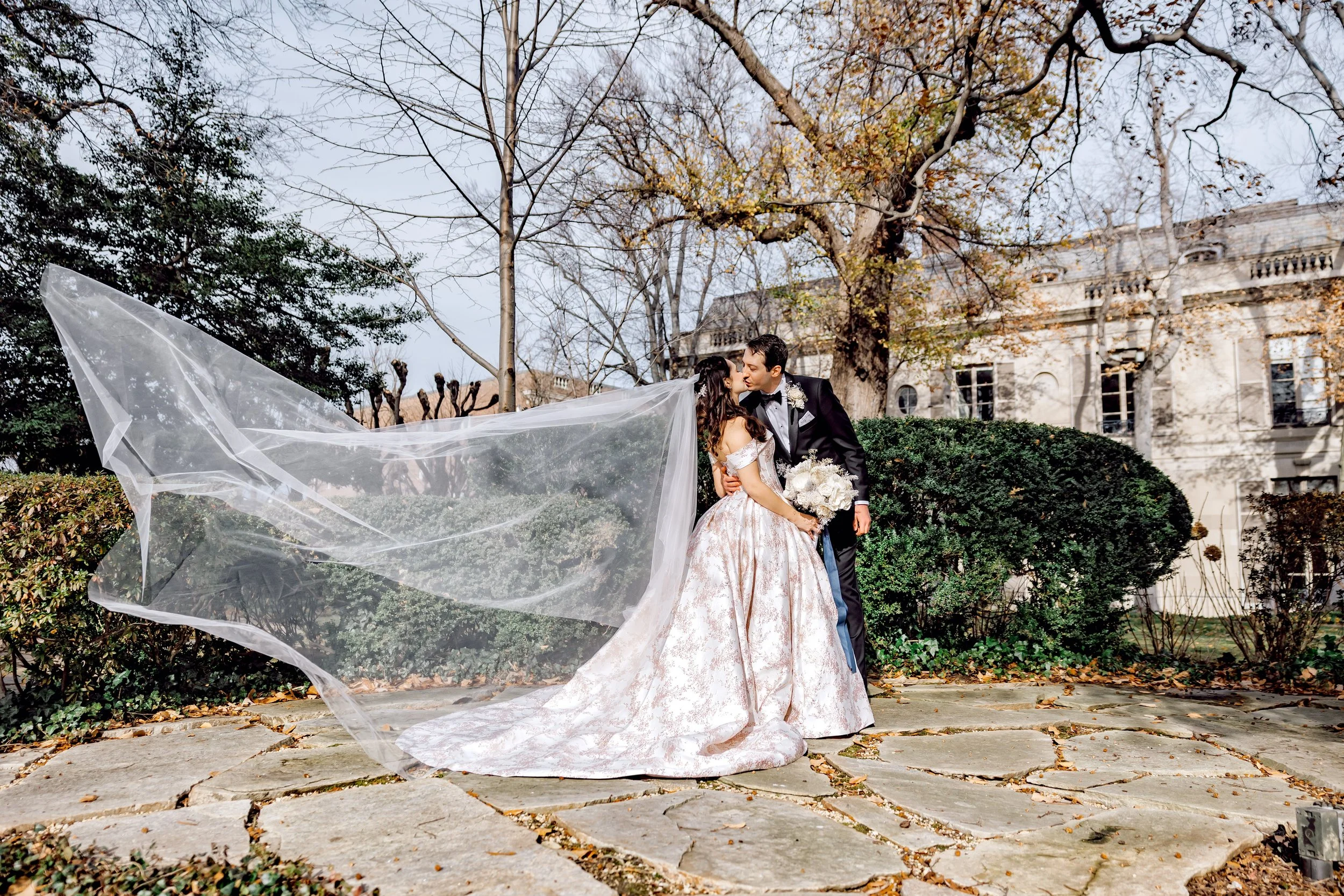 A newlywed couple sharing a kiss outdoors on a stone-paved area, with leafless trees and a historic building in the background. The bride wears a white gown with a long veil, and the groom is in a black tuxedo.