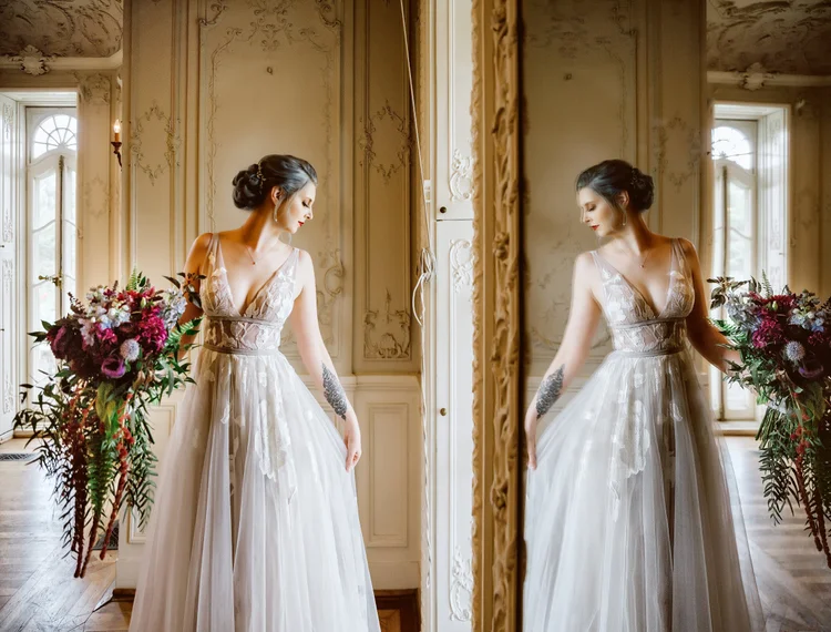 A woman in a wedding dress standing by a mirror, holding a large bouquet of flowers, in an ornate room with large windows and detailed wall decorations.