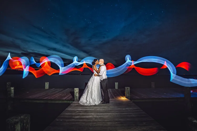 A couple in wedding attire kissing on a wooden dock at night, with colorful light streaks in the background over a dark sky and water.