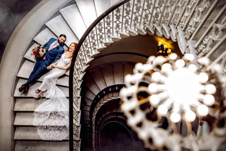 A bride and groom lying on a spiral staircase, looking up at the camera. The bride is in a white wedding dress and the groom is in a blue suit holding a bouquet of red and white flowers. The staircase has ornate white railings and is illuminated by a