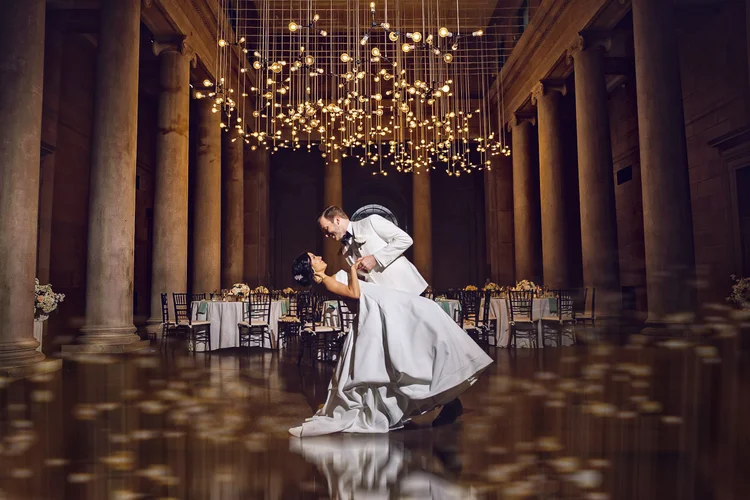 A bride and groom dancing at their wedding reception, with the groom in a white tuxedo and the bride in a white wedding gown, inside a grand hall with large columns and a chandelier overhead.