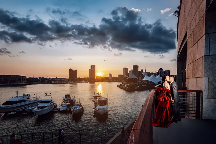 A couple kissing on a balcony overlooking a marina at sunset with city skyline in the background.