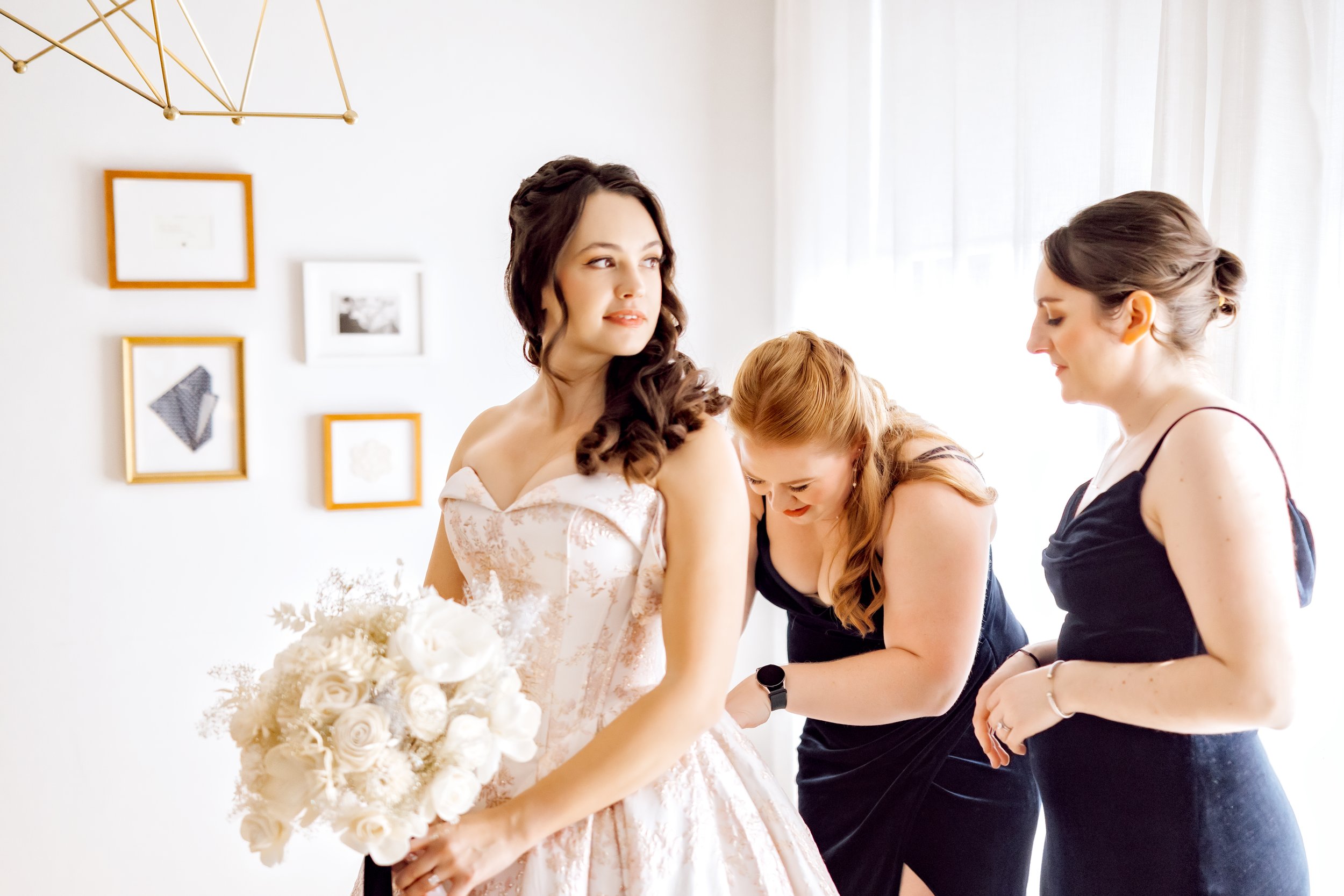 Bridal party getting ready, with a bride holding a bouquet of white roses and her bridesmaids assisting her in a well-lit room.