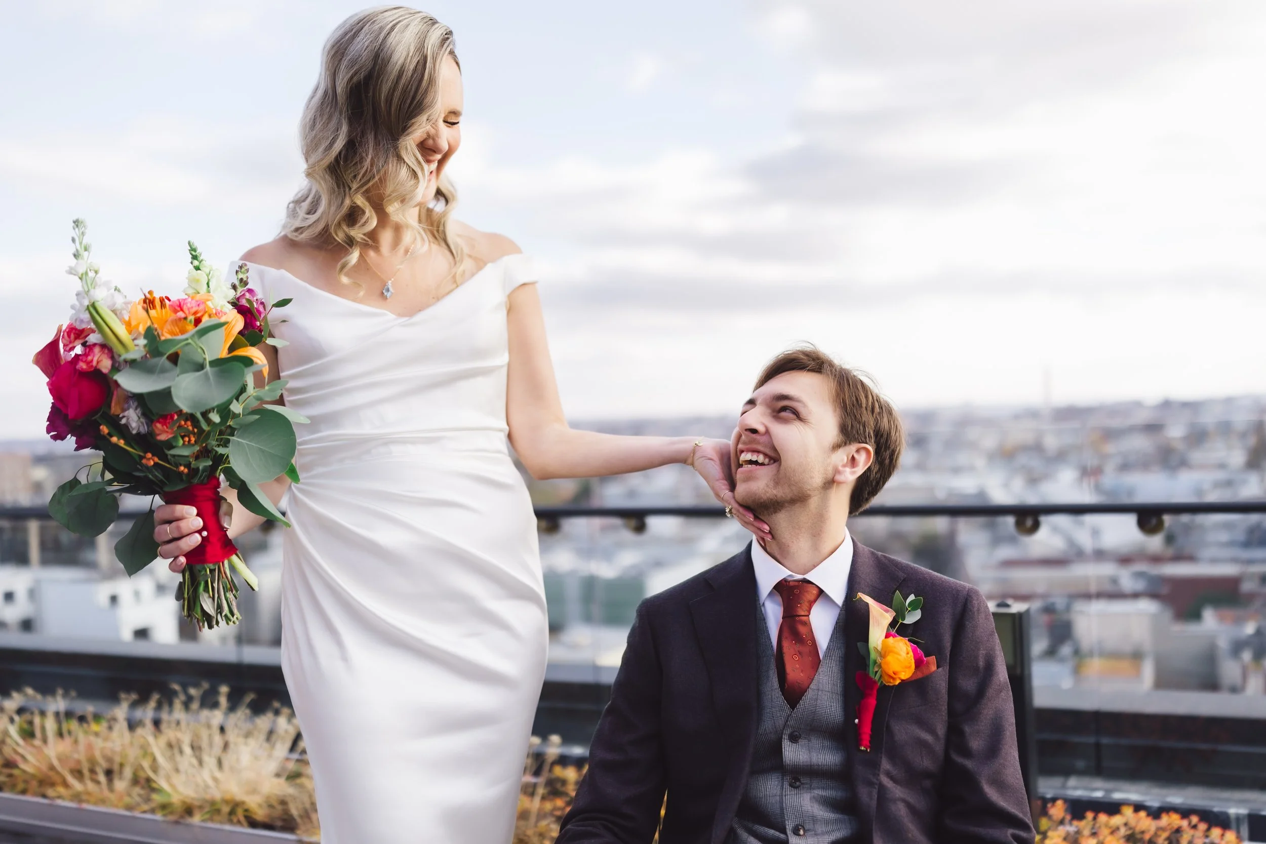 A woman in a white wedding dress holding a colorful bouquet of flowers, smiling at a man in a suit with a boutonniere, on a rooftop with cityscape in the background.