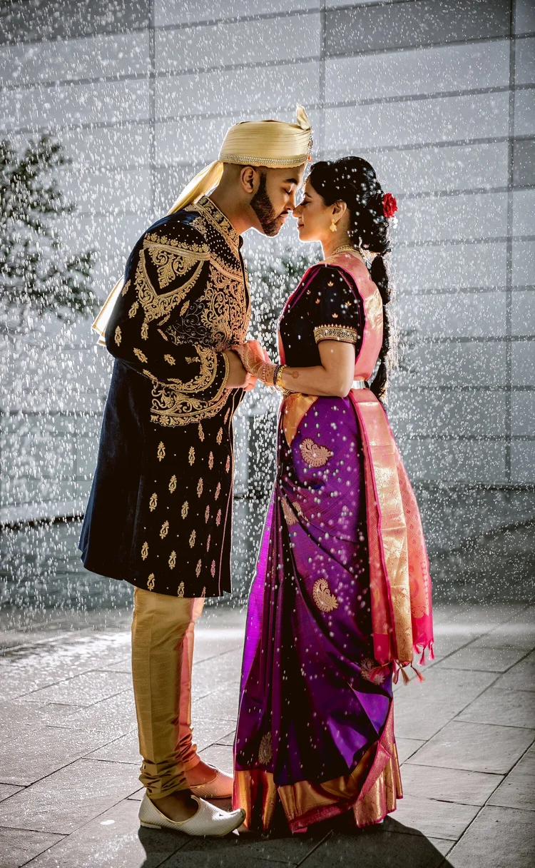 A couple in traditional Indian wedding attire stand close with foreheads touching in front of a water fountain, rain, or mist, with a modern building in the background.
